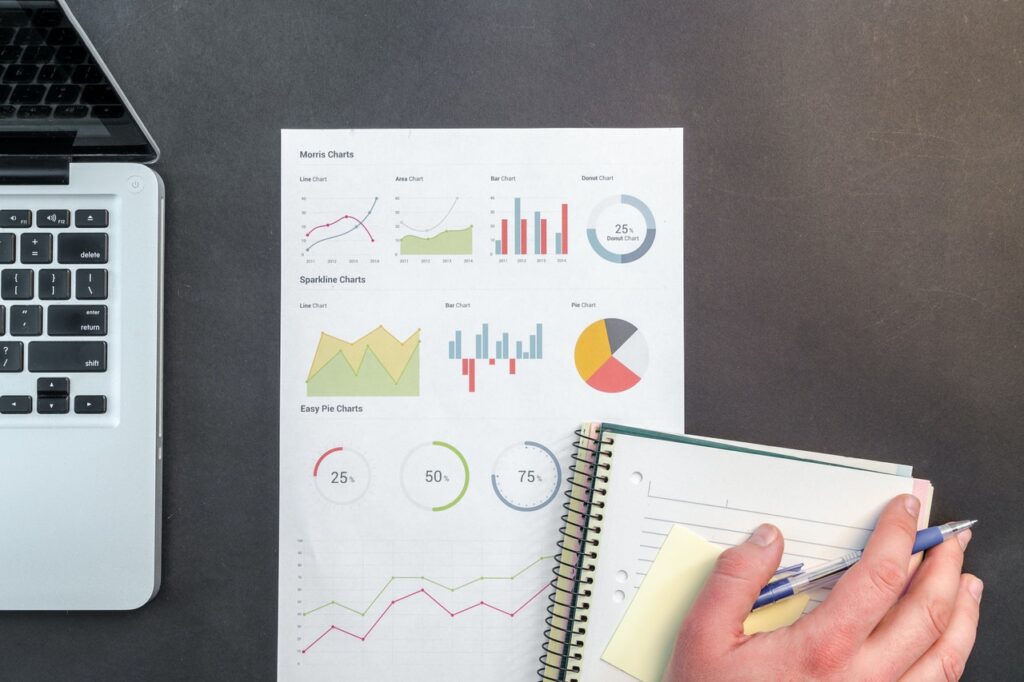 A desk scene with a laptop, printed analytics charts, and a hand writing notes in a notebook.