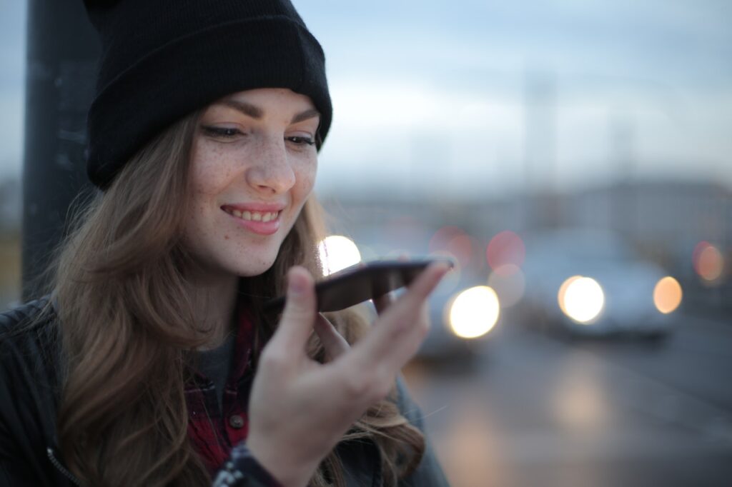 A woman wearing a black beanie is smiling while using her smartphone outdoors, with blurred car lights in the background.
